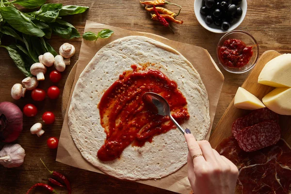 Abgeschnittene Ansicht einer Frau, die Tomatensauce auf Teig in der Nähe von Zutaten für Pizza auf einem Holztisch hinzufügt — Stockfoto