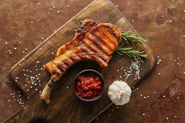 Blick von oben auf köstliches Ribeye-Steak mit Knoblauch und Tomatensauce auf Schneidebrett auf Steinoberfläche — Stockfoto