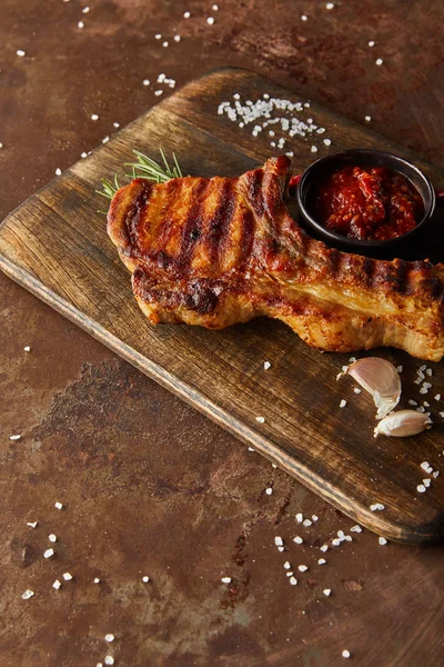 Vista de alto ángulo de delicioso bistec con ajo y salsa de tomate en tabla de cortar de madera en la superficie de piedra - foto de stock