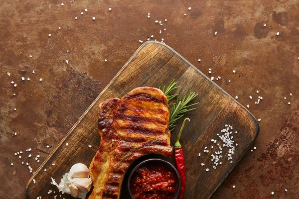 Draufsicht auf leckeres Steak mit Chili-Sauce, Knoblauch und Rosmarin auf Schneidebrett auf Steinhintergrund — Stockfoto