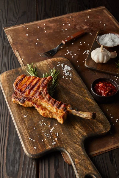 Focus selettivo della bistecca ribeye gourmet sul tagliere con sale, salsa di pomodoro e aglio su tavola di pietra su sfondo di legno — Foto stock