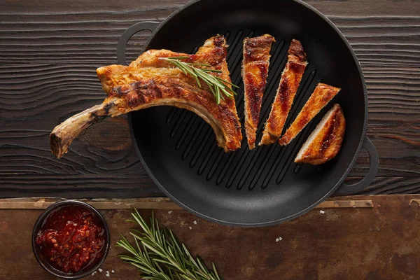 Top view of sliced ribeye steak in frying pan with tomato sauce and rosemary on wooden surface on stone background — Stock Photo