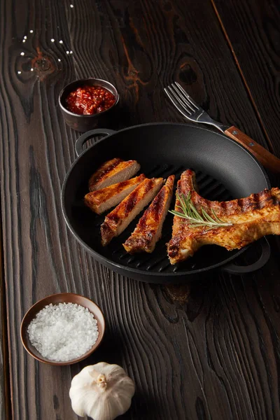 Selective focus of grilled steak in frying pan with tomato sauce, salt and garlic on wooden background — Stock Photo