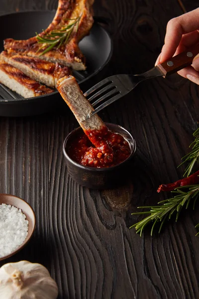 Cropped view of woman putting steak slice in tomato sauce by garlic and salt on wooden surface — Stock Photo