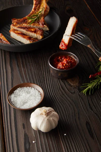 Selective focus of fork with steak slice by tomato sauce with salt and garlic on wooden surface — Stock Photo