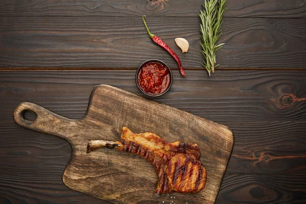 Blick von oben auf köstliches Ribeye-Steak auf Schneidebrett mit Chilisoße, Rosmarin und Knoblauch auf Holzgrund — Stockfoto