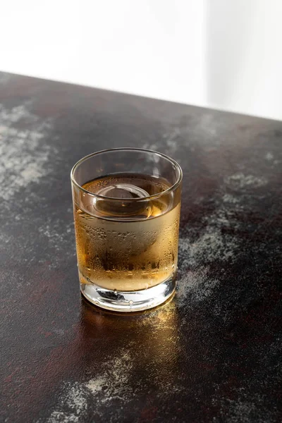 Wet glass with strong whiskey and ice cube on black surface — Stock Photo