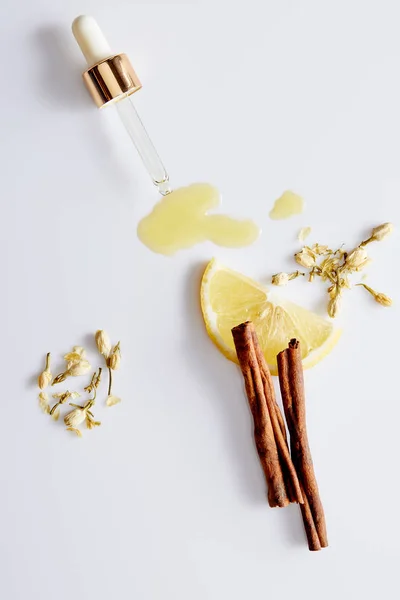 Vista superior del aceite cosmético que sale del gotero junto a rebanada de limón, palitos de canela y brotes de vainilla sobre fondo blanco - foto de stock