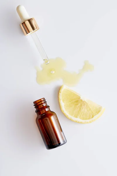 Vista dall'alto dell'olio cosmetico che fuoriesce dal contagocce accanto alla bottiglia e alla fetta di limone su sfondo bianco — Foto stock