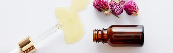 Vue de dessus de l'huile cosmétique qui coule du compte-gouttes à côté de la bouteille et des bourgeons roses sur fond blanc, prise de vue panoramique — Photo de stock