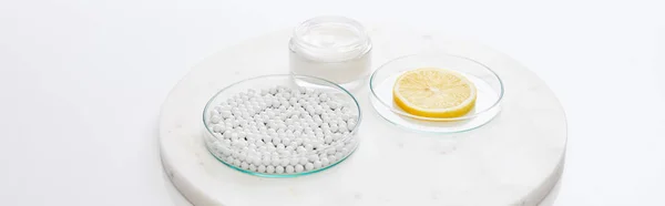 Laboratory glassware with decorative beads, slice of lemon next to cosmetic cream on round stand on white background, panoramic shot — Stock Photo