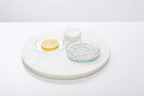 Artículos de vidrio de laboratorio con cuentas decorativas, rodaja de limón con crema cosmética en soporte redondo aislado en gris - foto de stock