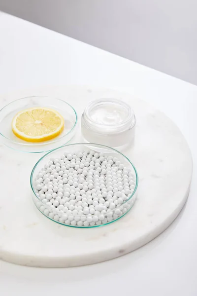 Laboratory glassware with decorative beads and slice of lemon next to cosmetic cream on round stand isolated on grey — Stock Photo