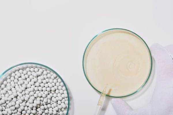 Cropped view of person holding laboratory glassware with sample of cosmetic cream and ampoule next to decorative beads on grey background — Stock Photo