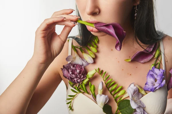 Vue recadrée de fille en soutien-gorge avec des fleurs violettes et violettes sur le corps tenant calla dans la bouche isolé sur blanc — Photo de stock