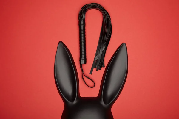 Vista dall'alto della maschera di coniglio nero e frusta fustigazione isolato sul rosso — Foto stock