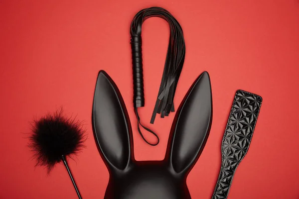 Vue de dessus du masque de lapin noir, de la pagaie, du chatouilleur de plumes et du fouet de flagellation isolés sur du rouge — Photo de stock