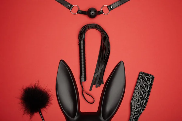 Top view of black rabbit mask, gag, paddle, feather tickler and flogging whip isolated on red — Stock Photo