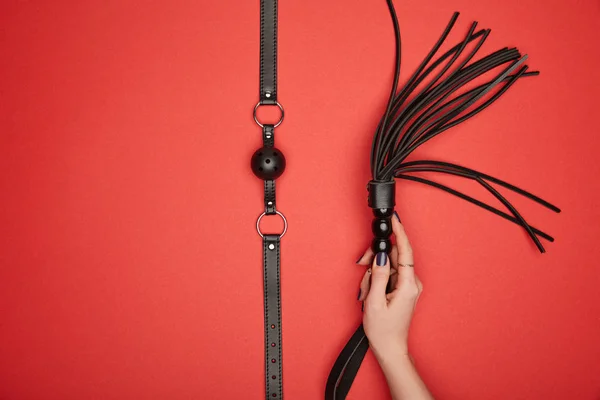 Cropped view of woman holding black flogging whip near gag isolated on red — Stock Photo