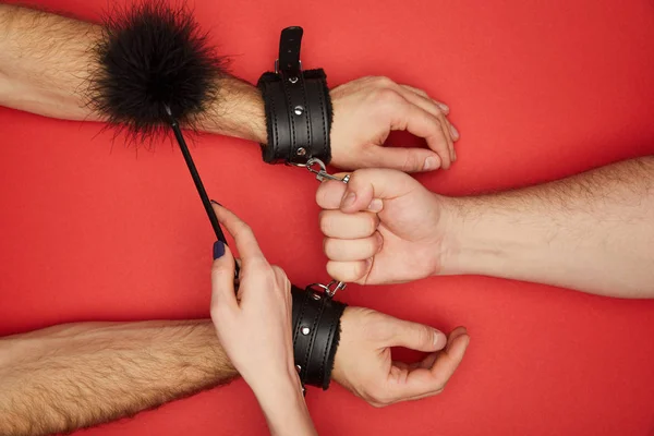 Cropped view of men and woman with handcuffs and feather tickler isolated on red — Stock Photo