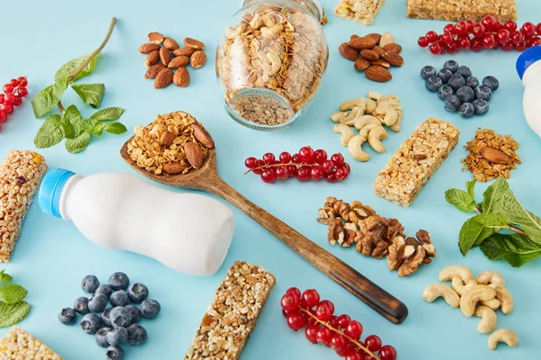 Frasco de granola, garrafas de iogurte, espátula de madeira e bagas com nozes em torno de fundo azul — Fotografia de Stock