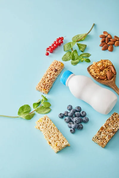 Botella de yogur, bayas, barritas de cereales, menta y almendras sobre fondo azul - foto de stock