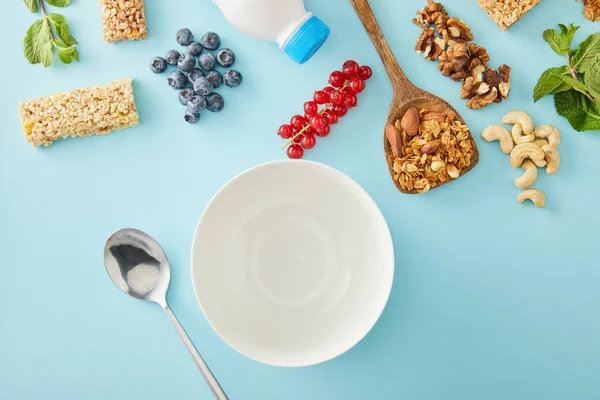Vista superior de cuenco vacío, cuchara, bayas, espátula, menta, nueces y barras de cereales sobre fondo azul - foto de stock