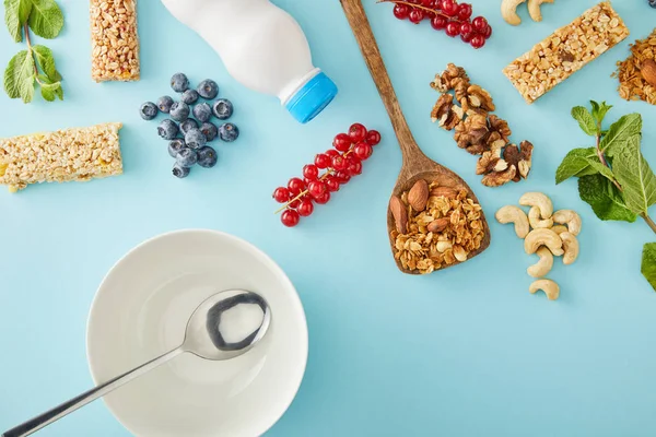 Vista superior de la cuchara dentro de un tazón vacío con bayas, menta, nueces, botella de yogur y barras de cereales sobre fondo azul - foto de stock