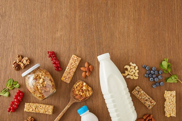 Vista superior de botellas de leche y yogur con bayas, menta, nueces, barras de cereales y tarro de granola sobre fondo de madera - foto de stock