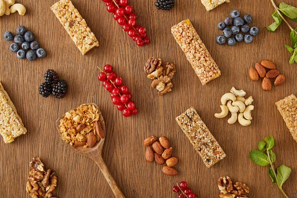 Top view of food composition of berries, nuts, cereal bars, mint and spatula with granola on wooden background — Stock Photo