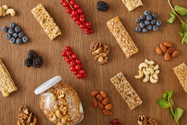 Top view of food composition of berries, nuts, cereal bars, mint and jar of granola on wooden background — Stock Photo