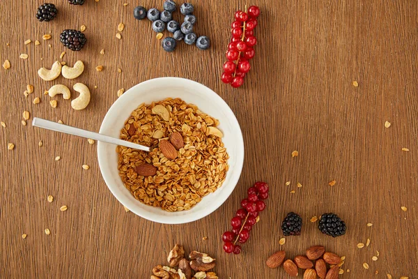 Top view of bowl with granola and blueberries, redcurrants, walnuts, almonds, cashews on wooden background — Stock Photo