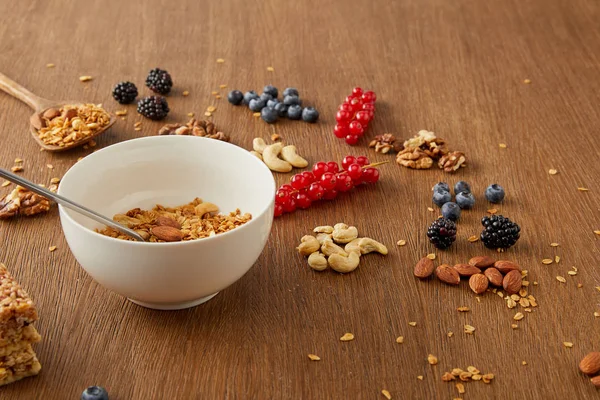 Bowl with granola next to blueberries, redcurrants, walnuts, almonds, cashews on wooden background — Stock Photo