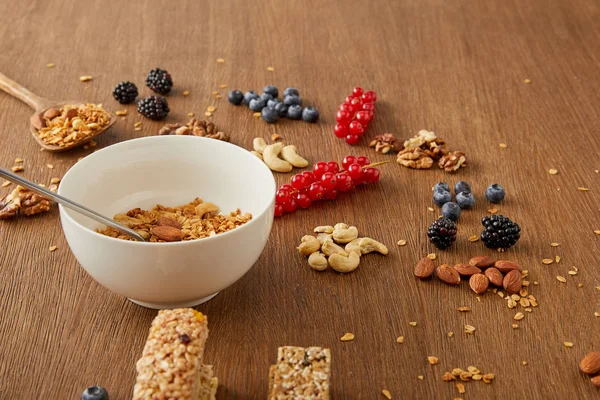 Cuenco con granola junto a bayas, frutos secos y barras de cereales sobre fondo de madera - foto de stock