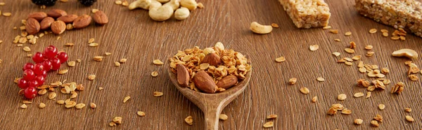 Espátula de madera con granola junto a grosellas rojas, nueces y barras de cereales sobre fondo de madera, plano panorámico - foto de stock