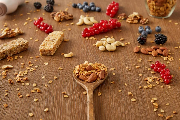 Holzspatel mit Müsli neben Beeren, Nüssen, Haferflocken und Müsliriegeln auf hölzernem Hintergrund — Stockfoto