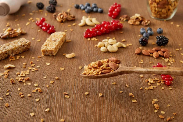 Espátula de madeira acima da mesa com granola ao lado de bagas, nozes e barras de cereais em fundo de madeira — Fotografia de Stock
