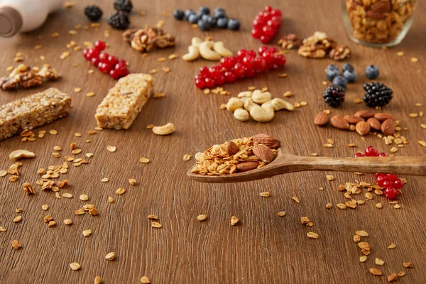 Wooden spatula above table with granola next to berries, nuts and cereal bars on wooden background — Stock Photo