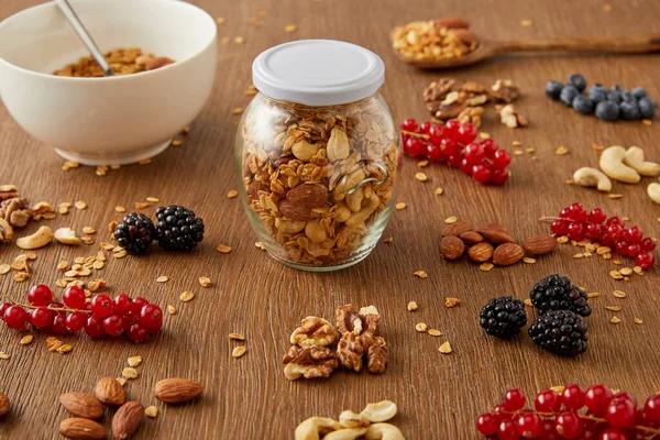 Selective focus of jar of granola and bowl next to nuts, oat flakes and berries on wooden background — Stock Photo