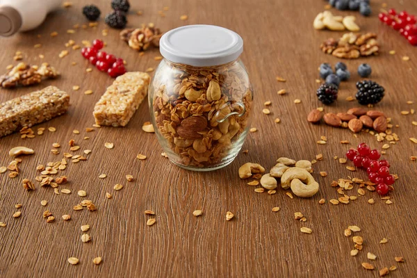 Foco seletivo de jarra de granola com nozes, flocos de aveia, bagas e barras de cereais em fundo de madeira — Fotografia de Stock