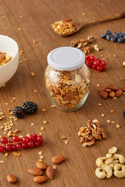 Jar of granola, bowl and spatula next to almonds, walnuts, cashews, berries on wooden background — Stock Photo