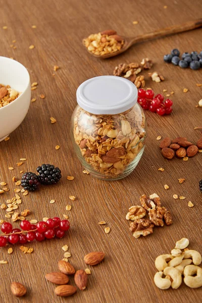 Tarro de granola, tazón y espátula junto a almendras, nueces, anacardos, bayas sobre fondo de madera - foto de stock