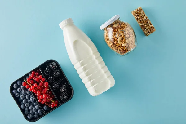 Vista superior del recipiente con bayas, botella de leche, tarro de granola y barra de cereales sobre fondo azul - foto de stock