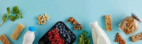 Top view of container with berries, bottles of yogurt and milk, jar of granola, nuts, cereal bars on blue background, panoramic shot — Stock Photo
