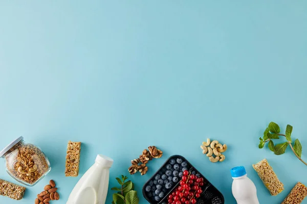Top view of container with berries, bottles of yogurt and milk, jar of granola, nuts, cereal bars and mint on blue background — Stock Photo