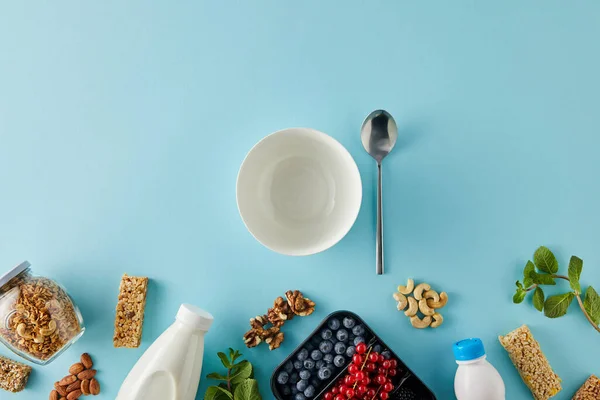 Vista superior do recipiente com bagas, garrafas de iogurte e leite, jarra de granola, nozes, barras de cereais, tigela, colher no fundo azul — Fotografia de Stock