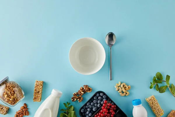 Draufsicht auf Container mit Beeren, Flaschen Joghurt und Milch, Glas Müsli, Nüsse, Müsliriegel, Schüssel, Löffel auf blauem Hintergrund — Stockfoto