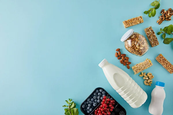 Vista superior del recipiente con bayas, botellas de yogur y leche, tarro de granola, nueces, barras de cereales sobre fondo azul — Stock Photo