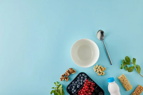 Верхний вид пустой миски с ложкой, контейнер с ягодами, бутылка йогурта, орехи, батончики, мята на синем фоне — стоковое фото