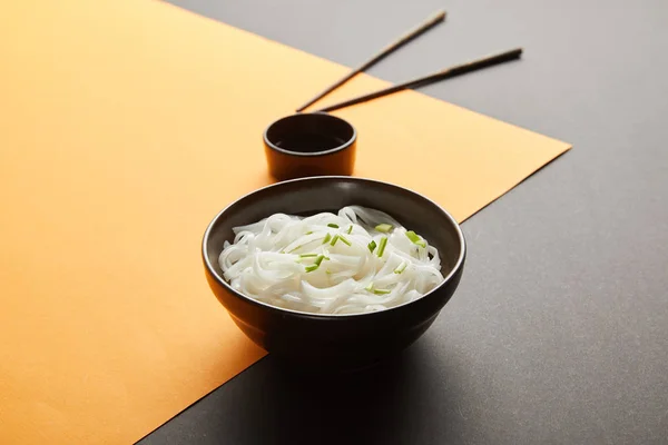 Selective focus of rice noodles in bowl near chopsticks and soy sauce on yellow and black surface — Stock Photo